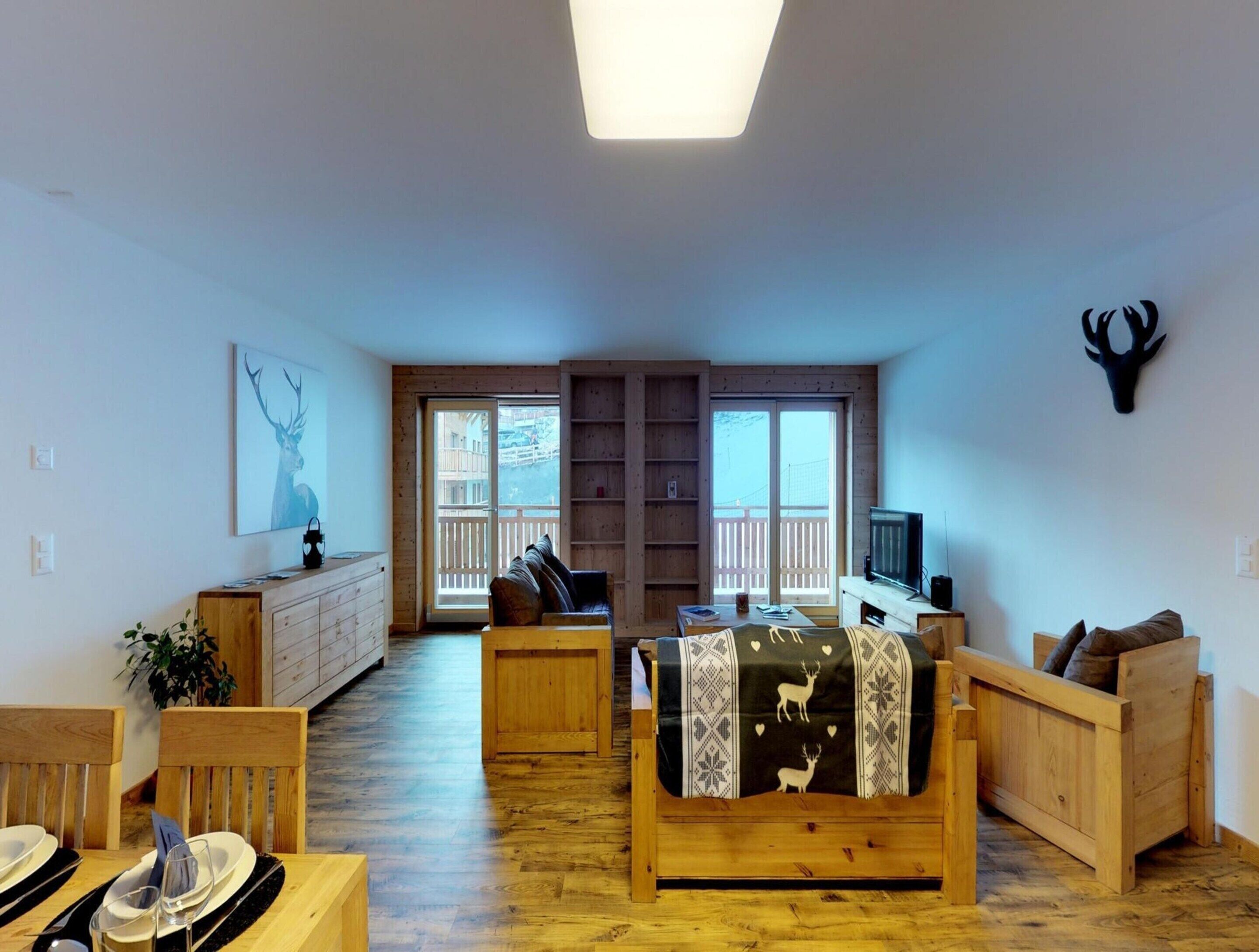Un salon chaleureux, doté de meubles en bois, d’un téléviseur et d’une grande fenêtre donnant sur un balcon.