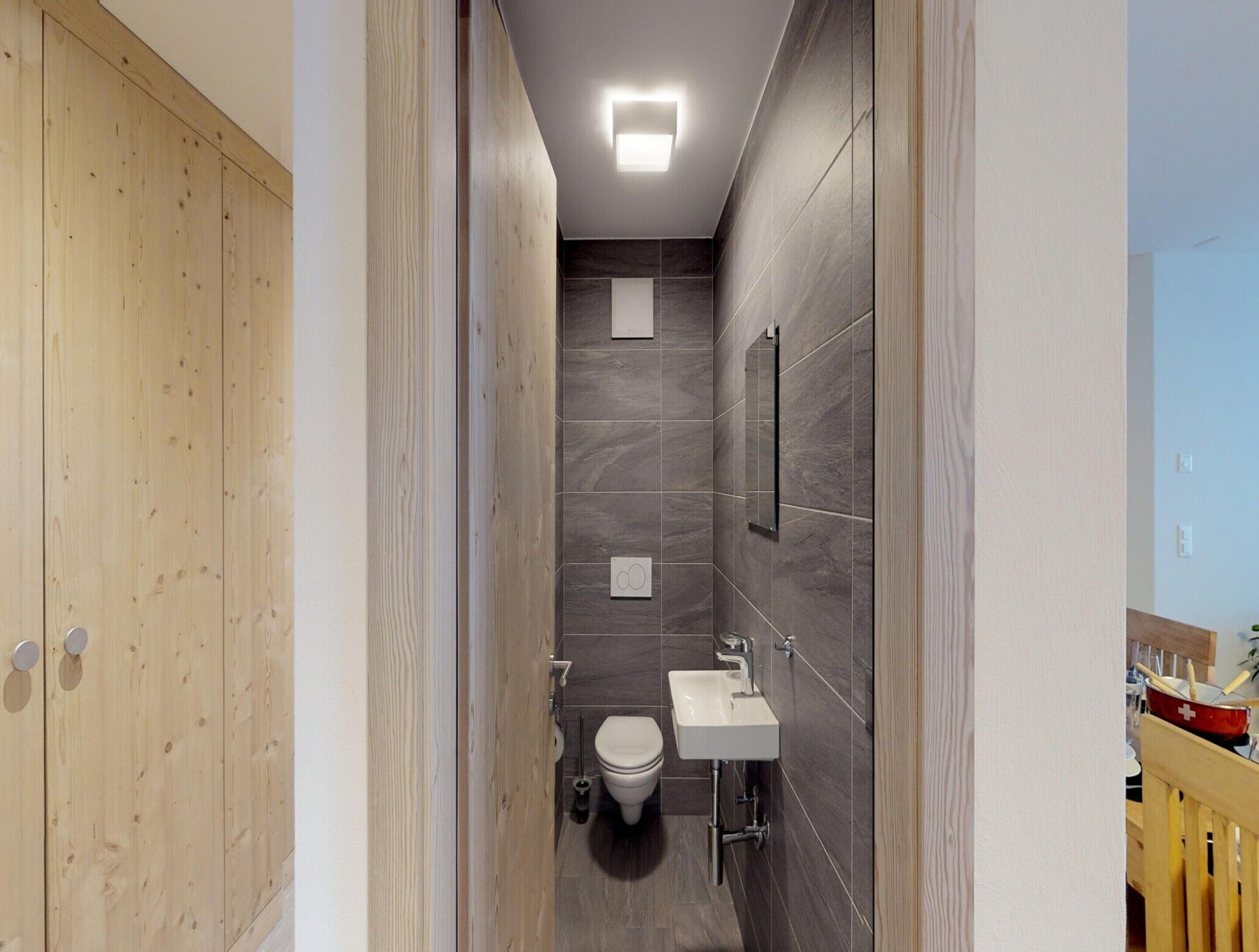 A narrow bathroom with a wooden door, a toilet, a sink, and a mirror on the wall.
