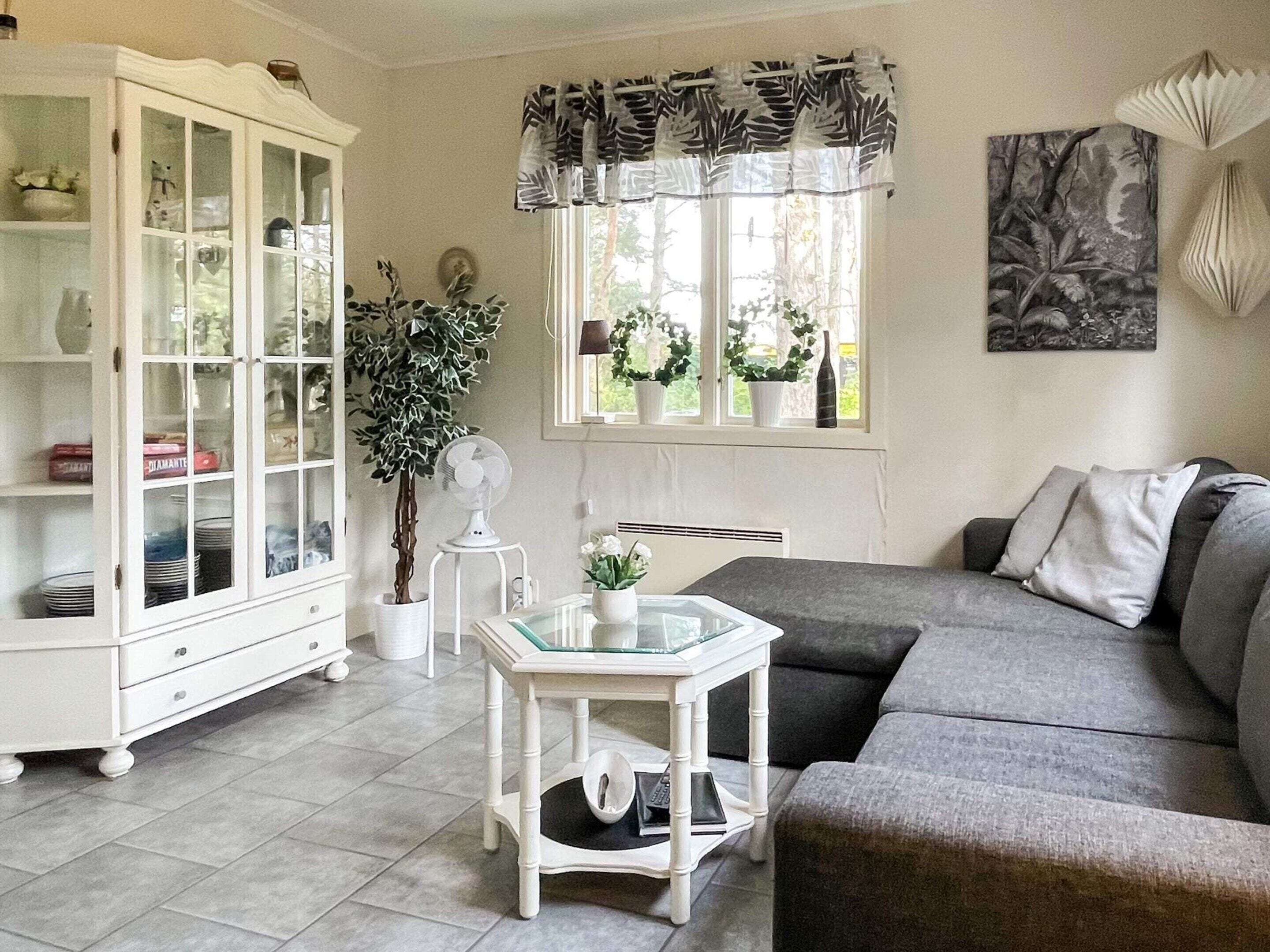 Una sala de estar con un sofá gris, un mueble blanco, una mesa de vidrio y una ventana con cortinas.