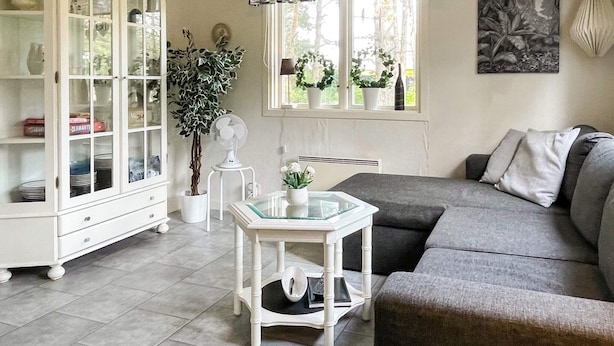 Una sala de estar con un sofá gris, un mueble blanco, una mesa de vidrio y una ventana con cortinas.