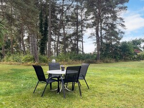 An outdoor dining area with a round table and four chairs, surrounded by a grassy field and tall trees.
