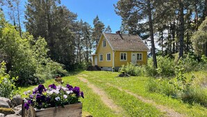 A yellow house with a sloped roof, surrounded by a forested area and a gravel path leading to it. - 4 Person Holiday Home in Farjestaden (Farjestaden)