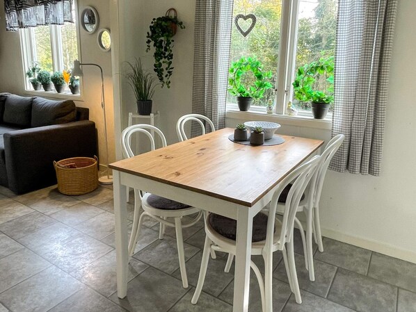 A dining area with a wooden table and white chairs, a window with curtains, and potted plants on the windowsill.