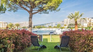 Vue d’une cour d’hôtel avec deux chaises longues, un grand arbre et, au fond, un port de plaisance.