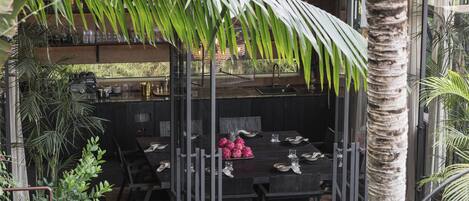 An outdoor dining area with a black table and chairs, surrounded by tropical plants and a palm tree.