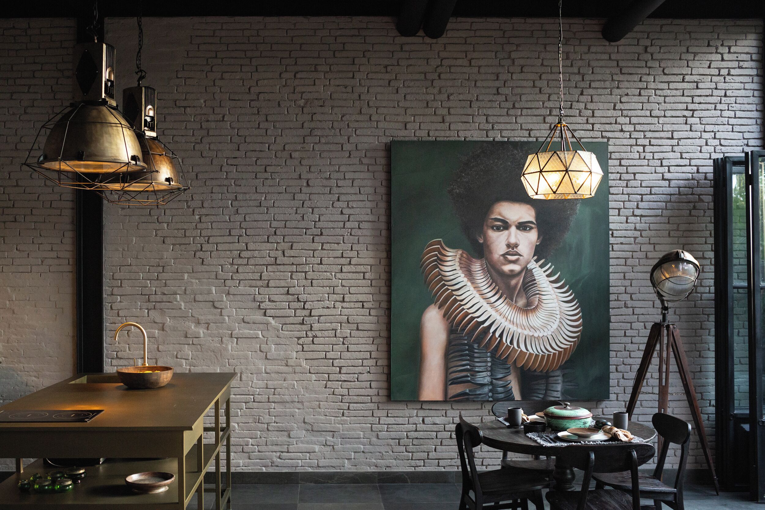 A dining area with a brick wall, a painting of a woman with a feathered collar, and pendant lighting.