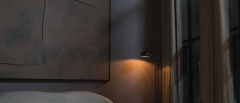 A bedroom with a bed, a wall-mounted abstract painting, a floor lamp, and a window with curtains.