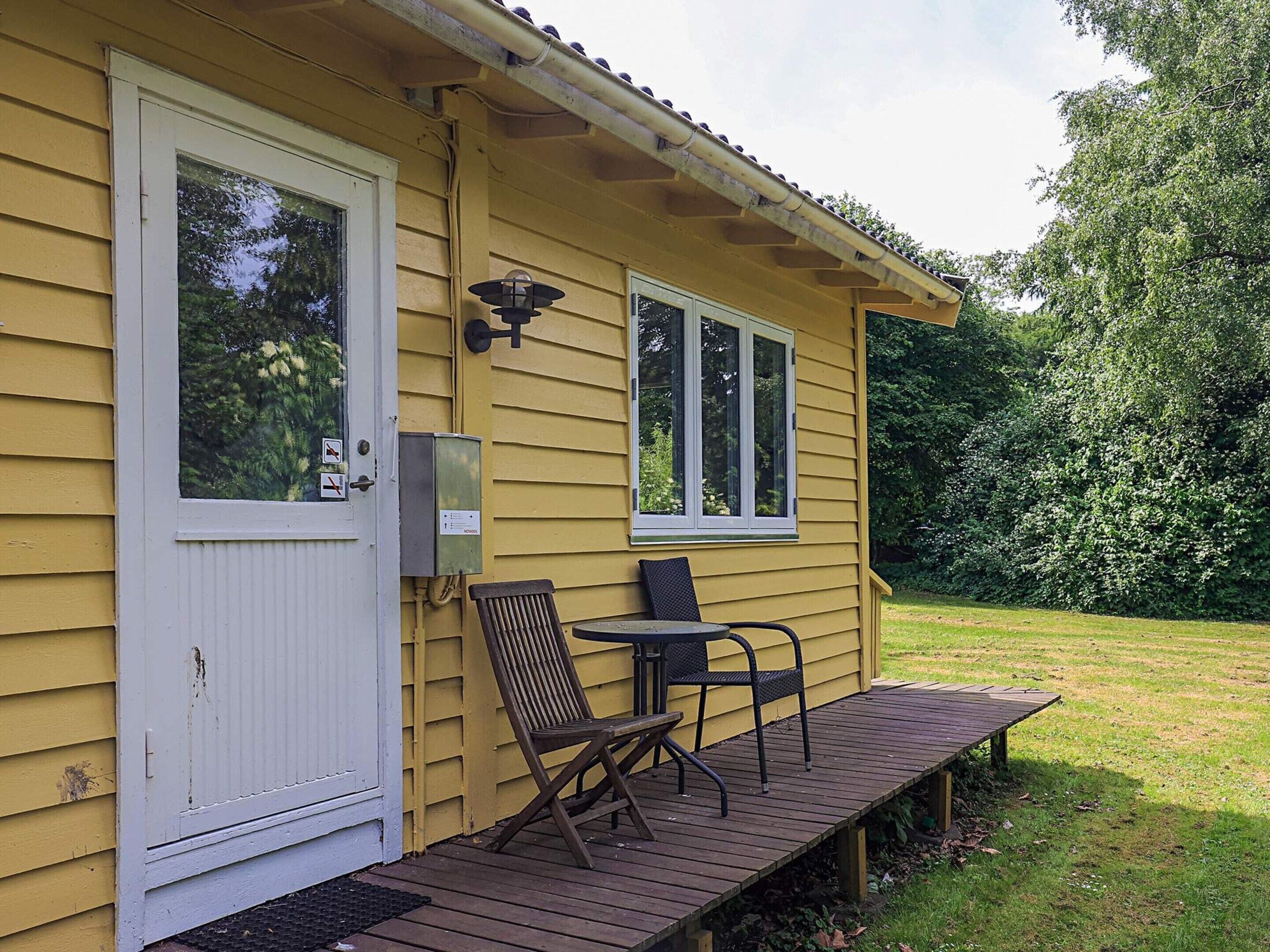 En gul byggnad med en vit dörr, två fönster, en veranda med ett bord och stolar samt en grön gräsmatta.