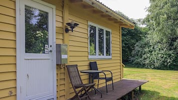 A yellow building with a white door, two windows, a porch with a table and chairs, and a green lawn.