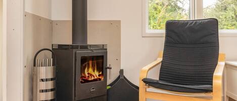 A room with a wood-burning stove, a chair, and a window with a view of trees.