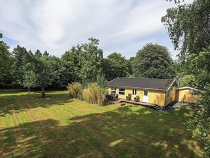 Une maison jaune d’un seul étage, dotée d’un patio et entourée d’une cour verdoyante et d’arbres.