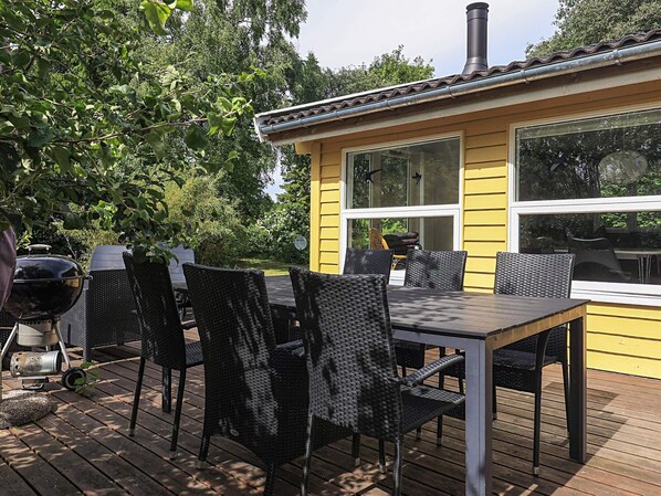 A deck with a dining table and chairs, a grill, and a yellow building with large windows.