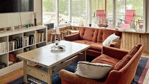 Un salon chaleureux avec une table en bois, un canapé et une bibliothèque. De grandes fenêtres, ornées de rideaux, laissent entrer la lumière naturelle et offrent une vue sur l’extérieur.