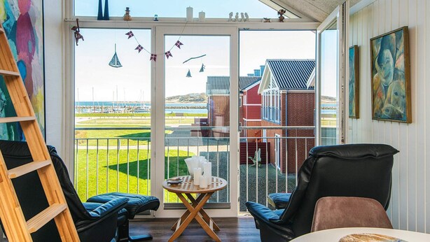 Een kamer met een grote glazen deur die uitkomt op een balkon met uitzicht op een jachthaven en gebouwen. Er staan twee stoelen, een klein rond tafeltje met een schaal, en een houten ladder die tegen de muur staat.