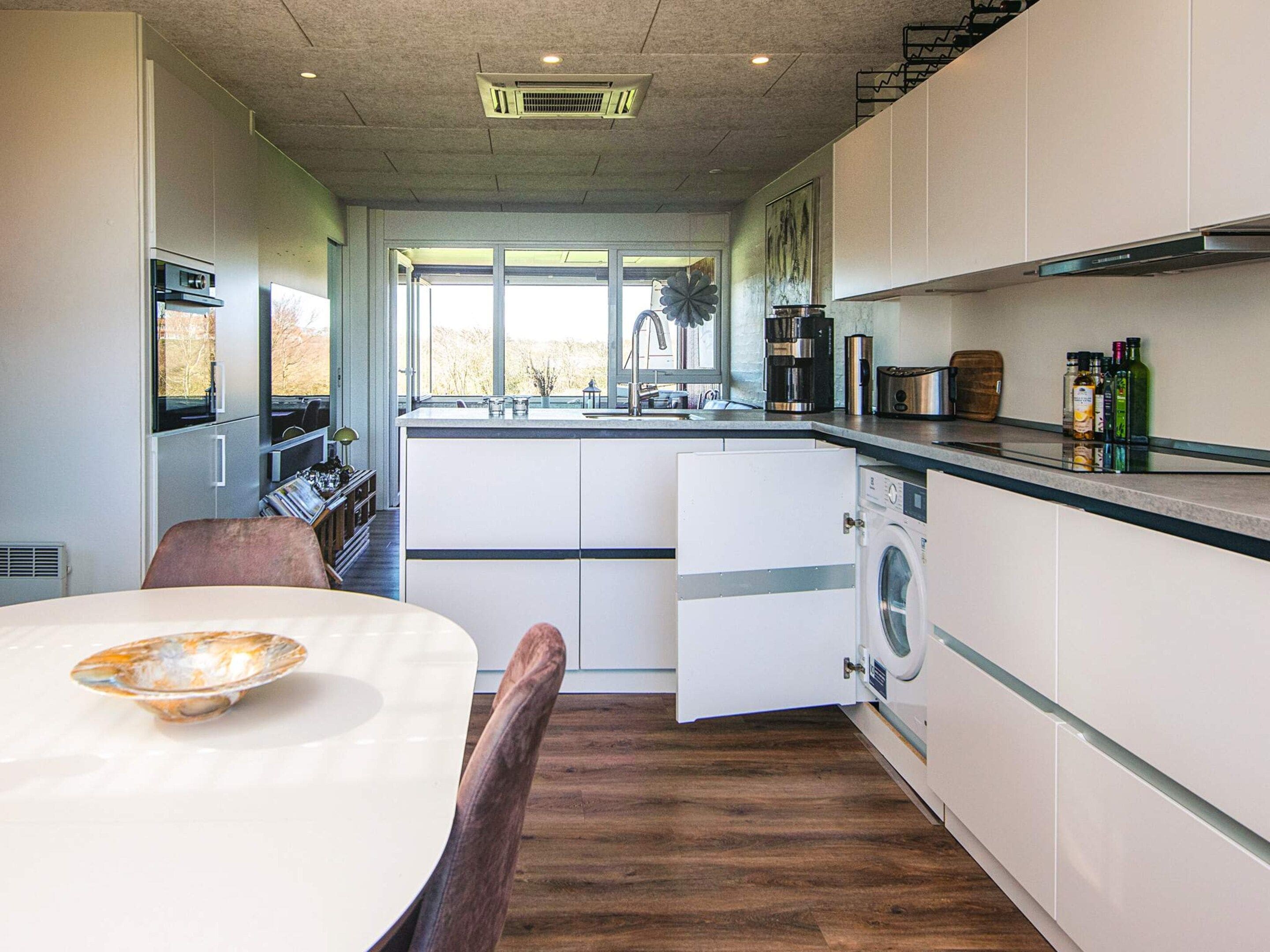 Een moderne keuken met een ingebouwde wasmachine, een eethoek met tafel en stoelen, en uitzicht door het raam.