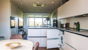 Een moderne keuken met een ingebouwde wasmachine en droger, een eethoek met een tafel en stoelen, en uitzicht op het omliggende landschap door grote ramen.