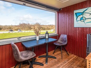 A room with a wooden floor, a red wall, a window with a view of a grassy area and parked cars, a table with two chairs, and a painting on the wall. - 4 Person Holiday Home in Lemvig (Lemvig)