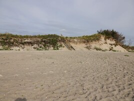 Una playa arenosa con una escalera de madera que sube hacia una duna.