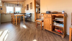 Ein gemütliches Zimmer mit einem Holzesstisch, Stühlen, einem Bücherregal mit Spielen und einem Fenster mit Vorhängen.