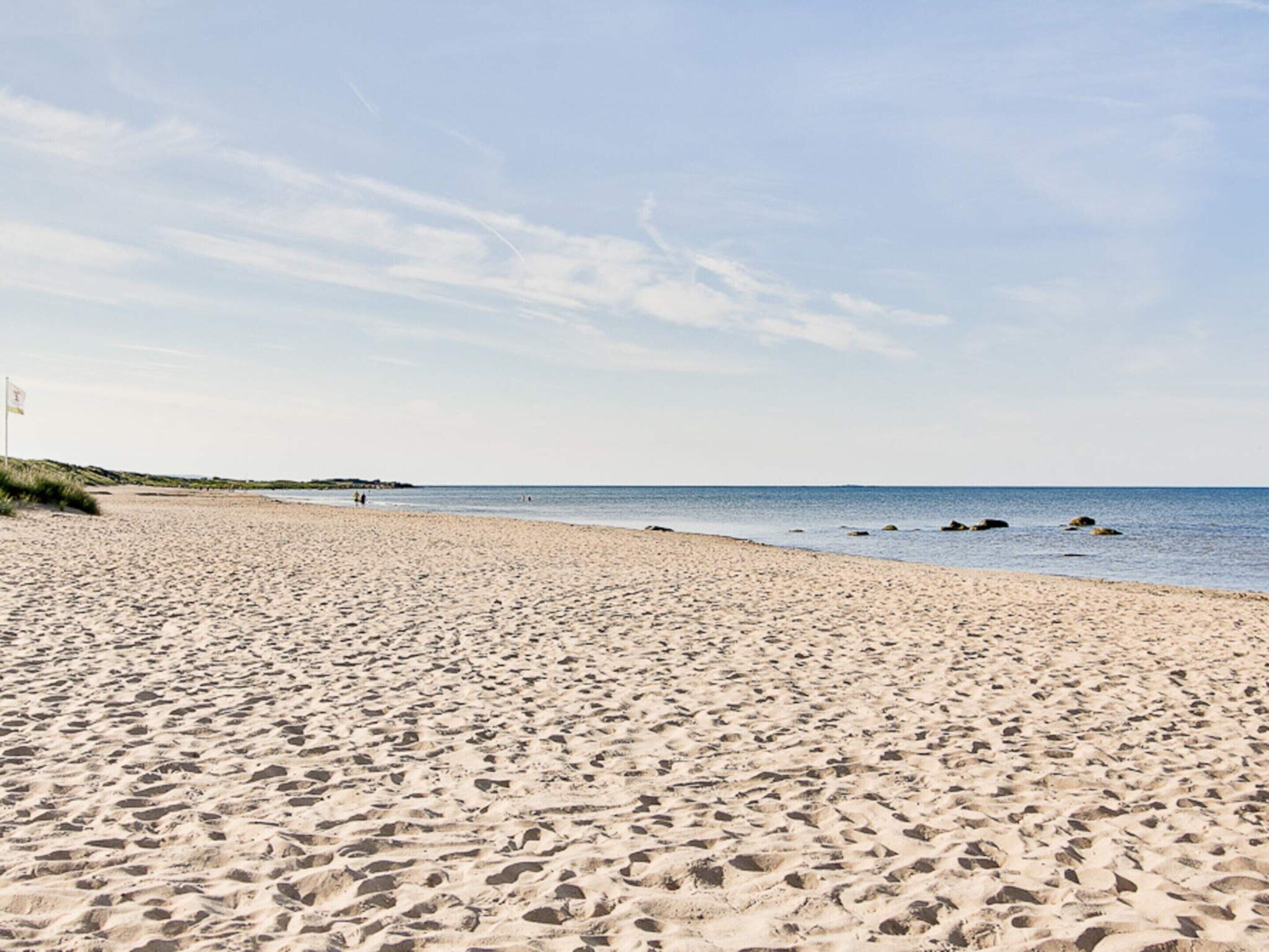 A wide sandy beach with footprints, a flag on the left, and calm water in the background.
