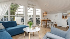 Een moderne woonkamer met een blauwe bank, een ronde salontafel en een grijze fauteuil. Er is een eethoek met een houten tafel en stoelen, en een keuken met witte apparaten.