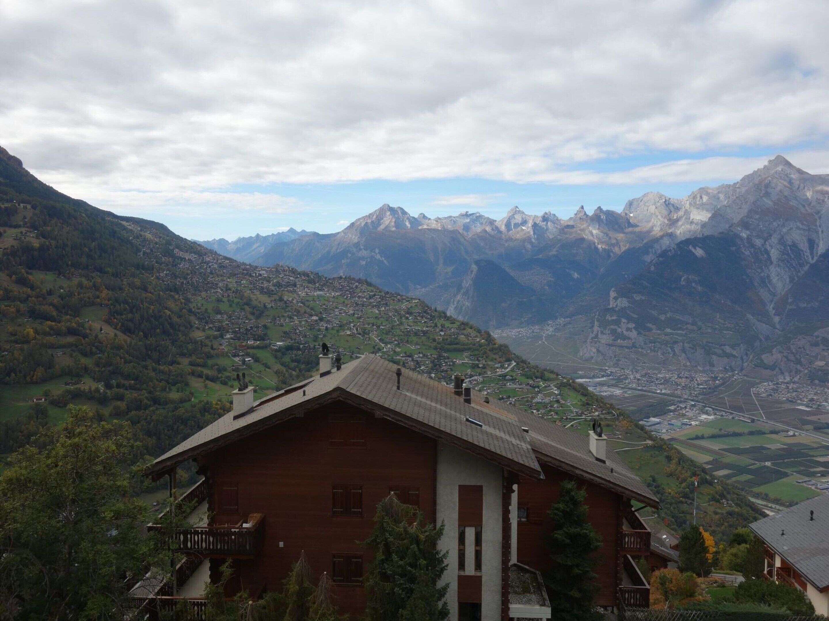 A wooden chalet with a balcony, set against a mountainous backdrop.