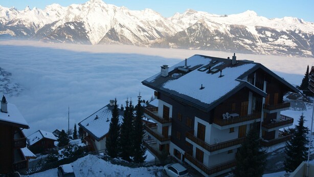 Ein mehrstöckiges Chalet mit schneebedecktem Dach, umgeben von schneebedeckten Bäumen und Bergen im Hintergrund.