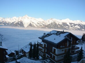 Un chalet de varios pisos con techo cubierto de nieve, rodeado de árboles nevados y montañas al fondo.