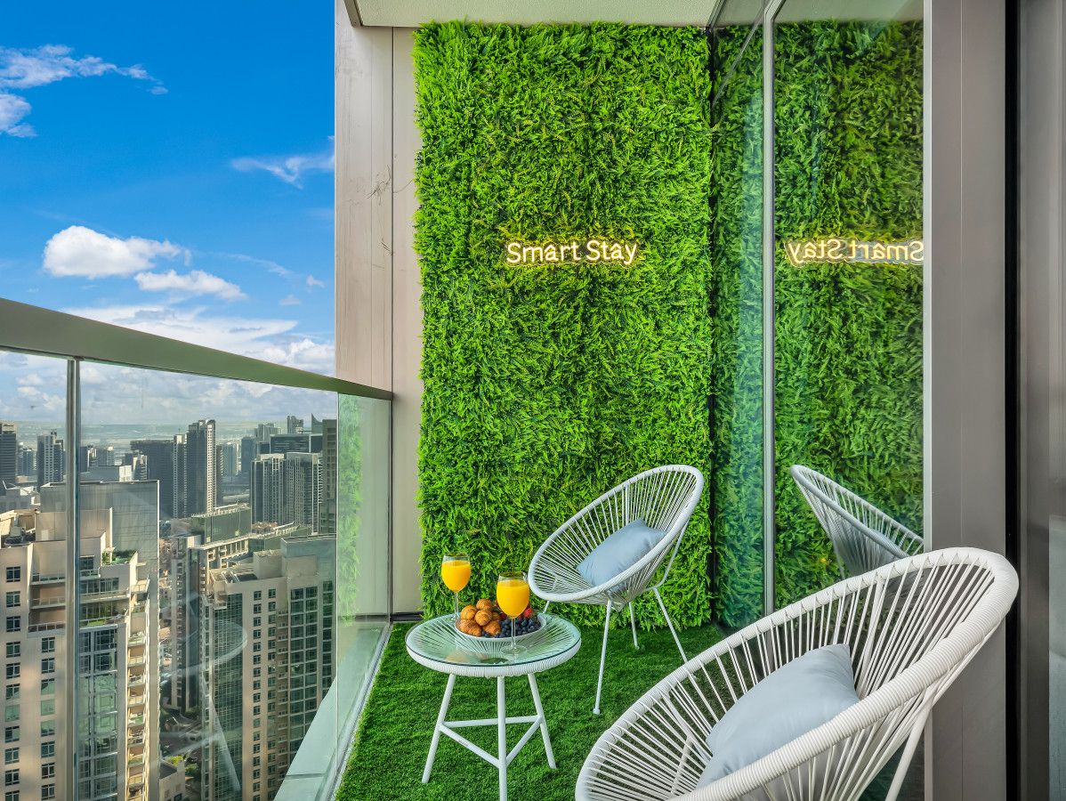 A balcony with a green wall, two white wicker chairs, a small table with fruit, and a city view.