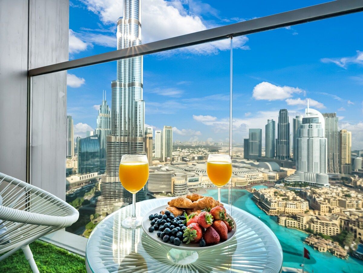 A balcony with a table set for breakfast, overlooking a cityscape with tall buildings and a clear sky.