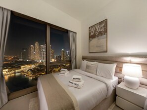 A hotel room with a bed, a nightstand, a lamp, a painting, and a view of a cityscape at night.