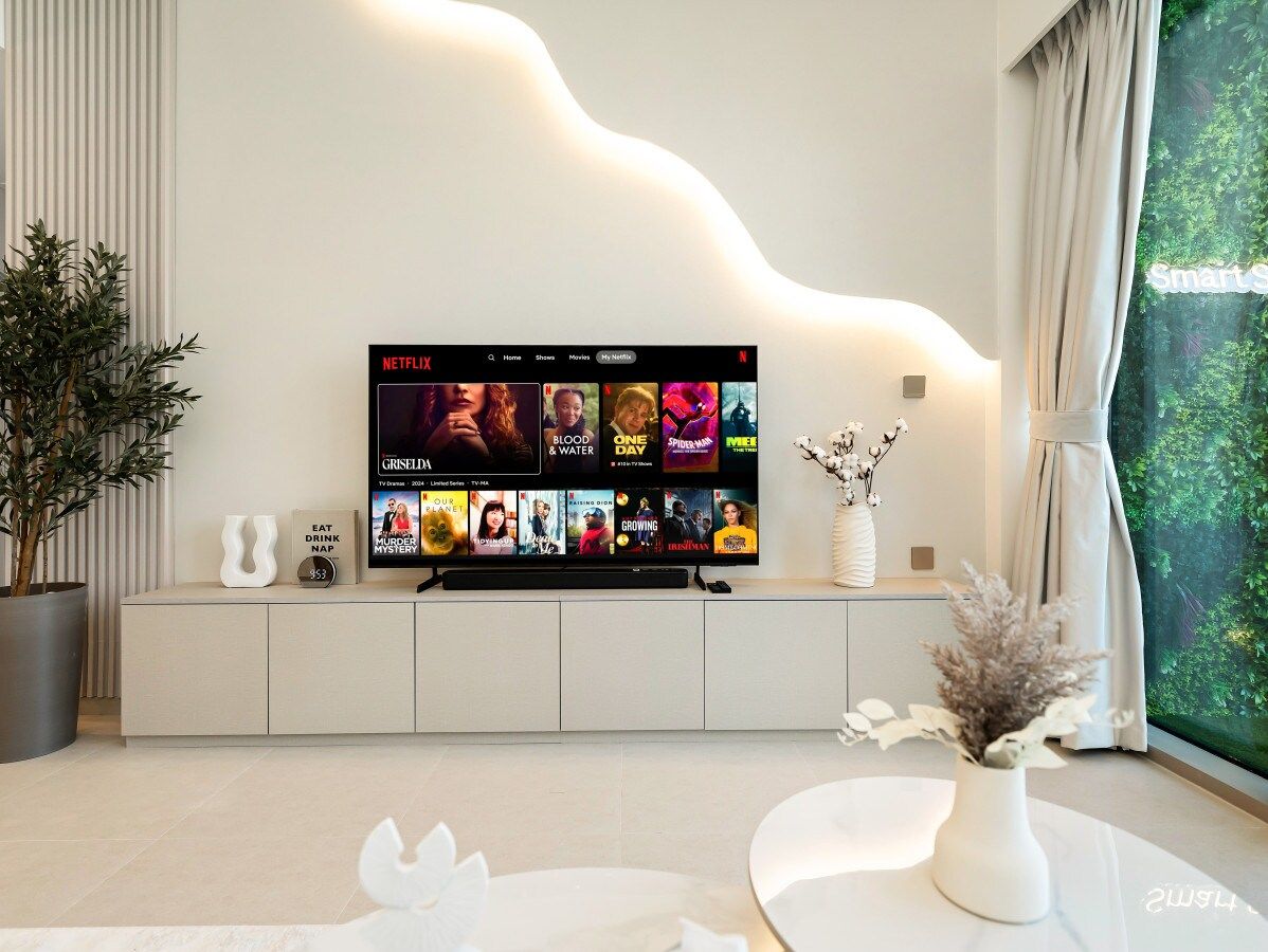 Ein modernes Wohnzimmer mit einem an einer weißen Kommode befestigten Flachbildfernseher, einer Topfpflanze und einer Vase mit getrockneten Blumen auf einem weißen Tisch.