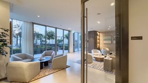 Una habitación de hotel moderna con un ventanal grande, un área de estar con sofás y una mesa de centro, y una mesita auxiliar.