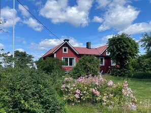 A red house with a sloped roof, surrounded by greenery and pink flowers. - 9 Person Holiday Home in Figeholm (Figeholm)