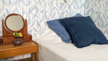 A neatly made bed with a white textured blanket and two blue pillows, a wooden nightstand with a round mirror and a vase of yellow flowers, and a wall with a leafy pattern.
