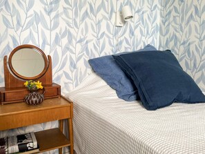 A neatly made bed with a white textured blanket and two blue pillows, a wooden nightstand with a round mirror and a vase of yellow flowers, and a wall with a leafy pattern. - 9 Person Holiday Home in Figeholm (Figeholm)