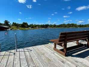 A wooden dock with a bench, a buoy, and a calm lake with boats and houses in the background.