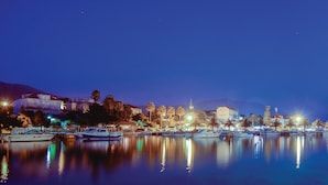 A marina at dusk with boats docked, a church with a lit tower, and buildings with lights.