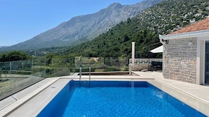 A swimming pool with a glass railing, a stone wall, and a view of mountains.