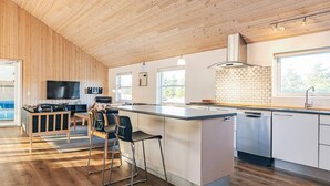 Et moderne køkken med træloft, en central ø, barstole, et fjernsyn og et stort vindue.