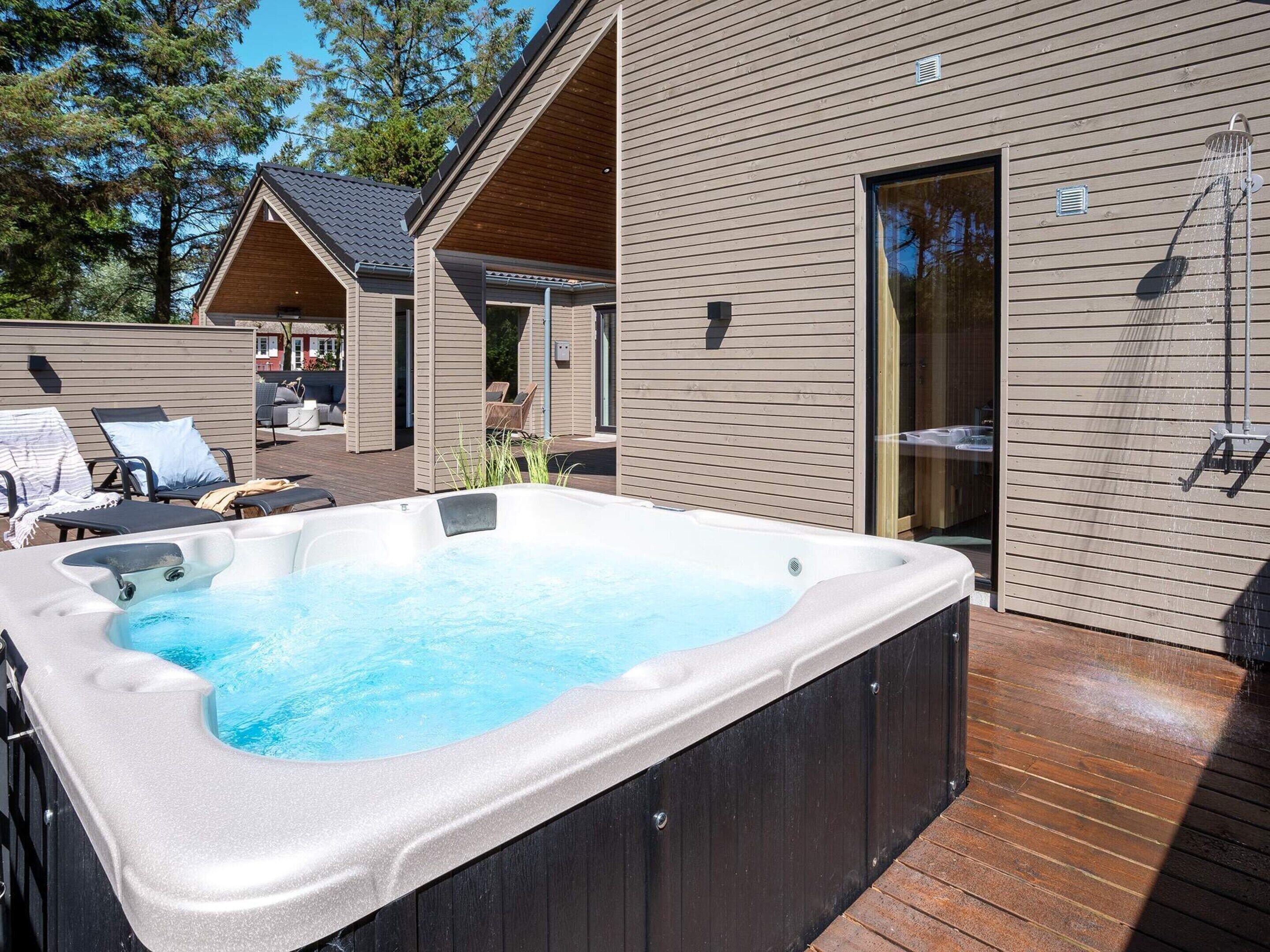 Un bain à remous sur une terrasse en bois, avec vue sur une maison moderne et un espace salon extérieur abrité.