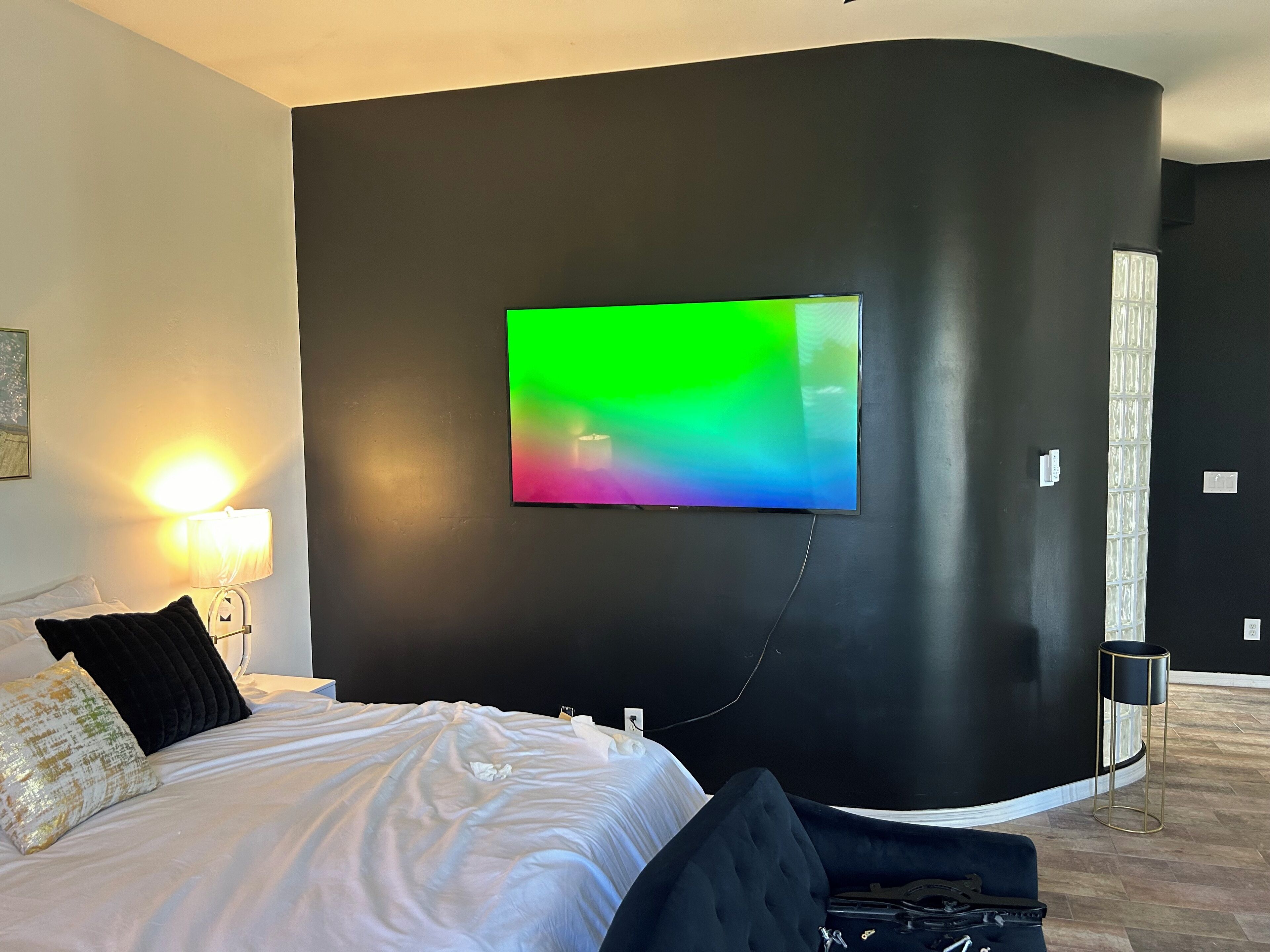 Una habitación de hotel moderna con un televisor de pantalla plana grande montado en una pared oscura, una cama con sábanas blancas y almohadas negras, una lámpara de noche y una pequeña mesita de noche negra.