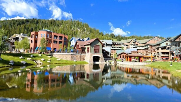 A lakeside hotel with a reflection in the water, surrounded by buildings and a forested mountain backdrop.