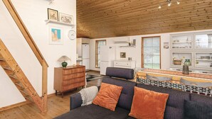 A modern living room with a wooden ceiling, a sofa with orange cushions, a wooden dresser, and a kitchen area with white cabinets and a window. - 4 Star Holiday Home in Hovborg (Hovborg)
