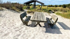 A wooden picnic table with benches on a sandy beach, a wooden shelter in the background, and a clear blue sky. - 5 Person Holiday Home in Borgholm (Borgholm)