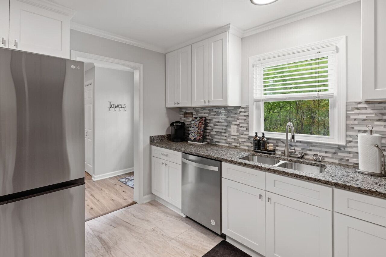 A modern kitchen with white cabinetry, a stainless steel refrigerator, a granite countertop, and a window with blinds.