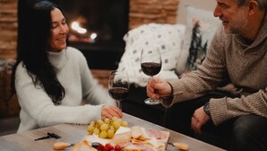 A couple seated at a table with a charcuterie board and wine glasses, in a cozy room with a fireplace in the background.