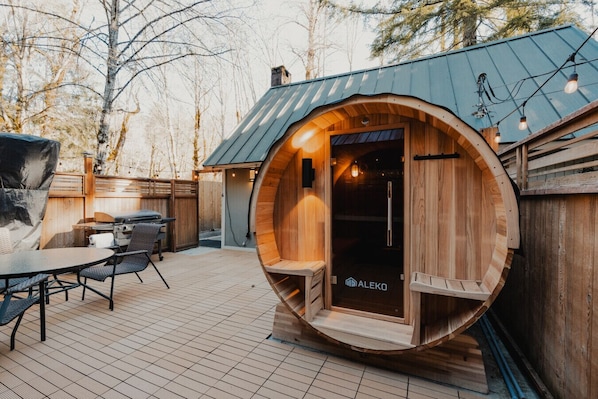 A wooden sauna with a curved exterior, a small patio area with a table and chairs, and a metal roof.