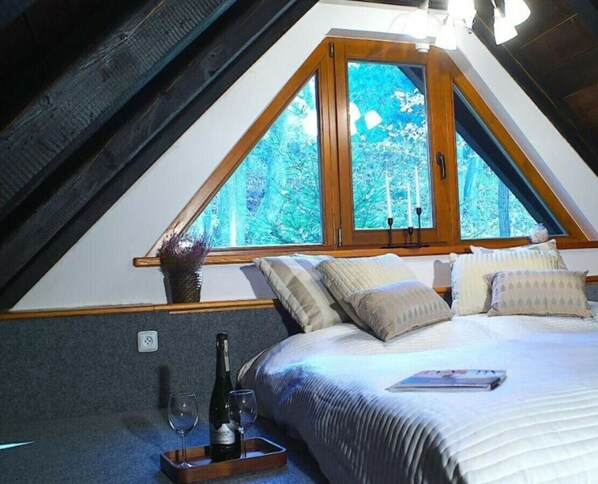 A bedroom with a sloped ceiling, a large window with a view of trees, a bed with pillows, a bedside table with a bottle and glasses, and a vase with flowers. - Comfortable Villa With Facilities (Benecko)