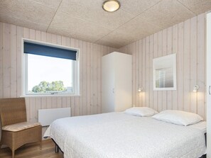 A bedroom with a bed, a chair, a wardrobe, a window, and a wall-mounted light fixture.